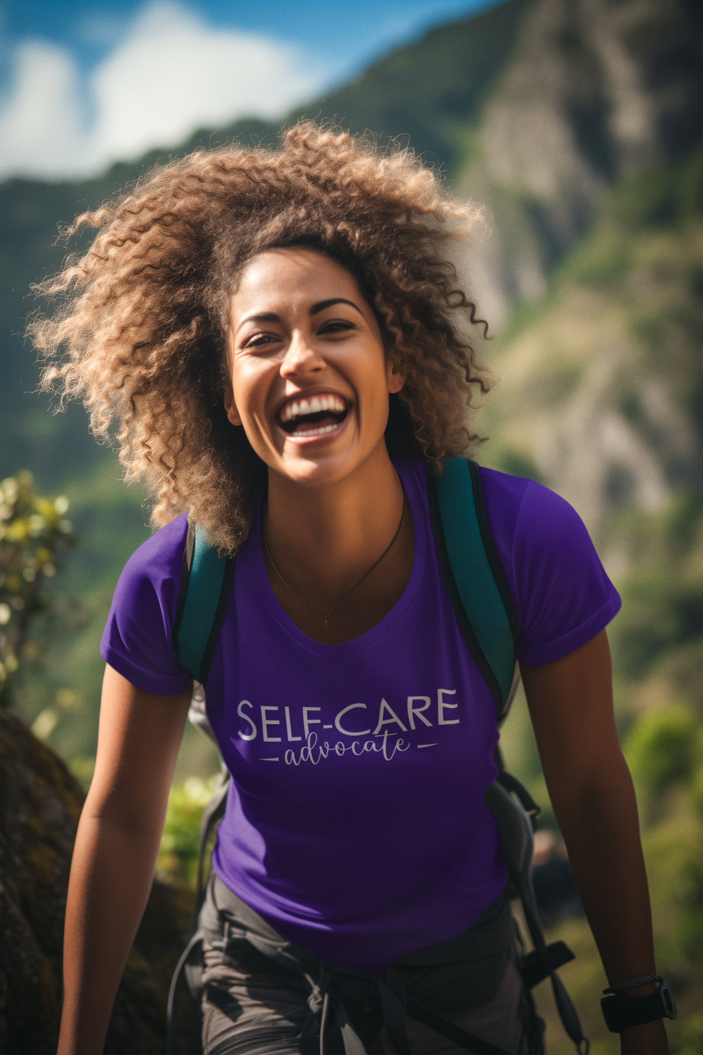 A person with curly hair is smiling, wearing a purple "Self-Care Advocate Tee" from Sharp Tact Kreativ—crafted from 100% ring-spun cotton in unisex sizing—and a backpack, standing outdoors with mountains in the background.