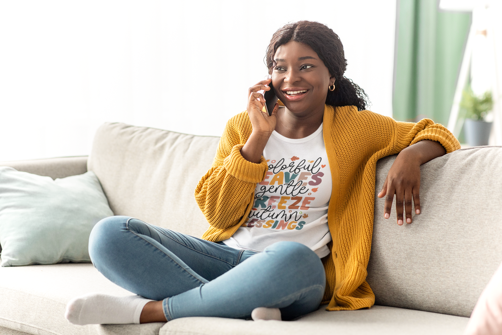 Sitting cross-legged on the sofa, a woman wearing the "Colorful Leaves, Gentle Breeze, Autumn Blessings Tee" by Sharp Tact Kreativ chats joyfully on her cellphone. Her vibrant outfit perfectly complements her surroundings, making it seem as though she’s just stepped out of an afternoon with nature lovers.