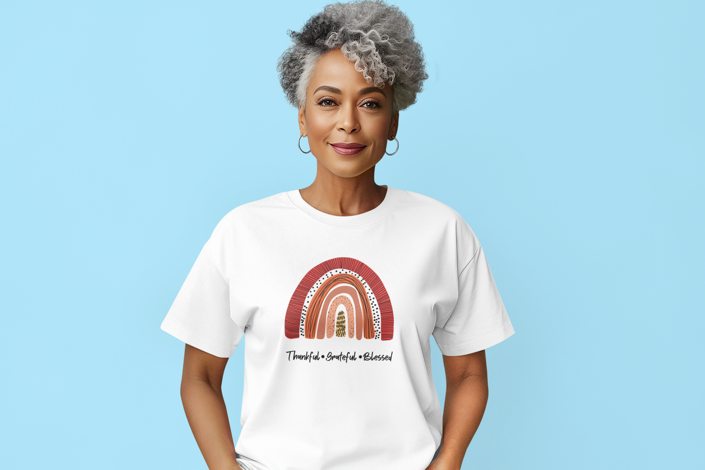 A person with short gray curly hair stands against a blue background, wearing the Sharp Tact Kreativ "Thankful, Grateful, Blessed Tee," a white ringspun cotton T-shirt featuring a colorful rainbow design and uplifting text.