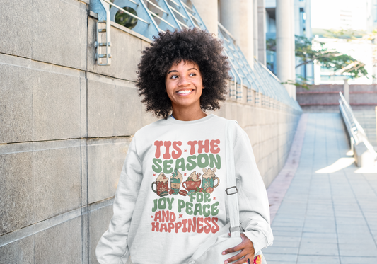 A person with curly hair smiles while wearing the Sharp Tact Kreativ cozy crewneck sweatshirt in unisex sizing, featuring a festive design that reads, "Tis the Season for Joy, Peace, and Happiness," as they stand outdoors near a stone building. The cotton polyester blend provides comfort and warmth in the chilly air.